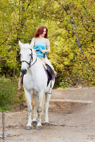 Young smiling red-haired woman rides on white horse in park