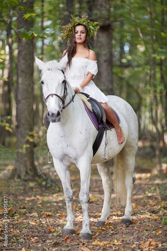 Barefooted girl in white dress with floral wreath on head sits on horseback in park