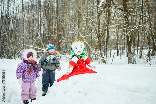 Little boy and girl stand looking at stuffed dummy Maslenitsa that sits on snowbank in winter park