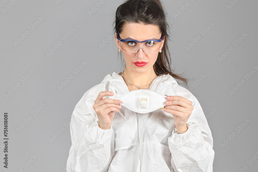 Half-growth portrait of woman in white protective suit of synthetic paper and glasses, looking at us, holds respiritory half mask in hands, on gray background Half-growth portrait of woman in white protective suit of synthetic paper and glasses, looking at us, holds respiritory half mask in hands, on gray background