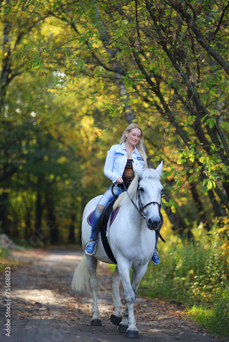Blonde woman rides on white horse on path in sunny autumn park