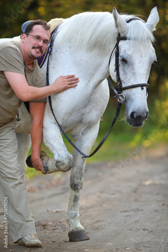 Happy man holds hoof of beautiful white horse in sunny autumn park