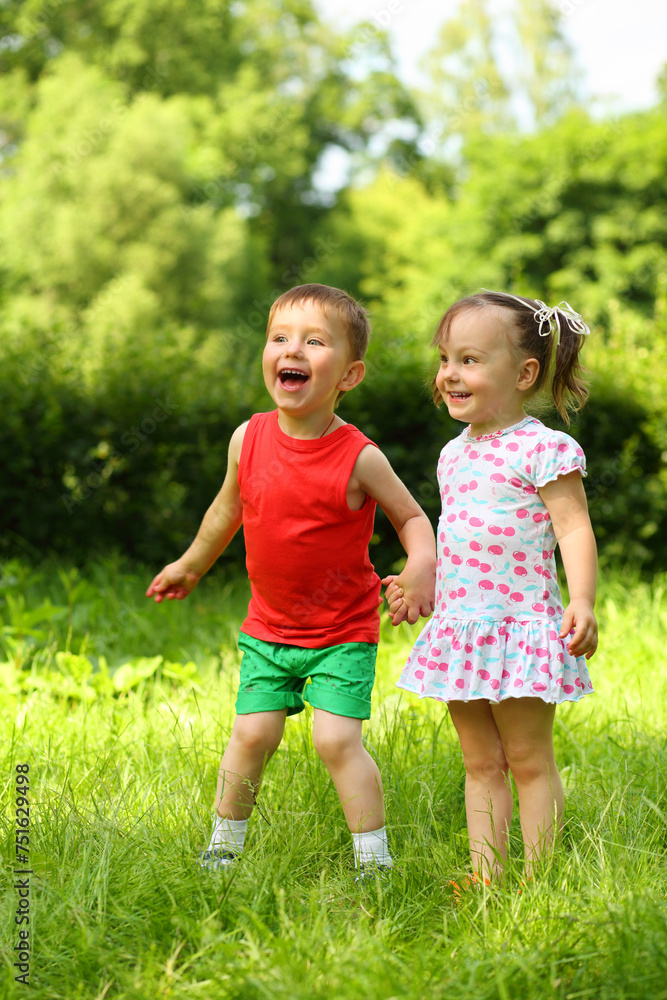 Little laughing girl and boy stand on fresh green grass in park