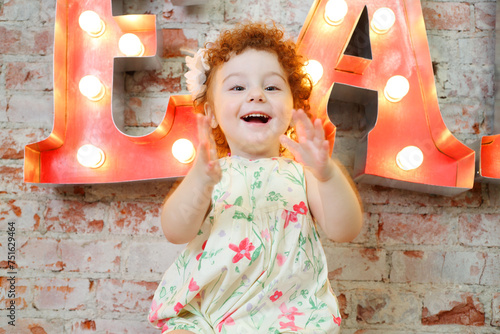 Little happy girl claps and sits near wall with big letters in studio photo