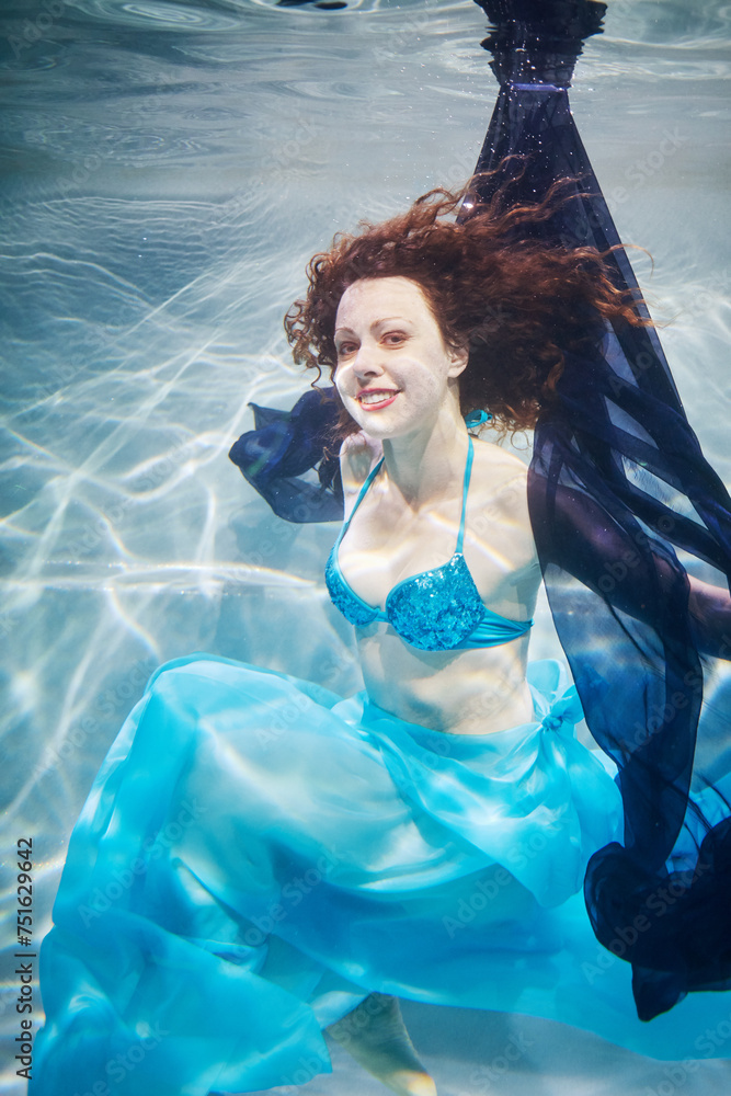 Smiling young woman in blue swimsuit poses in swimming pool with dark-blue fabric underwater - obrazy, fototapety, plakaty 