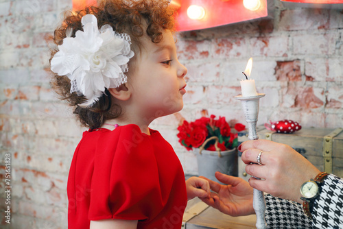 Little cute girl in red blows out white candle in mother hands in room photo