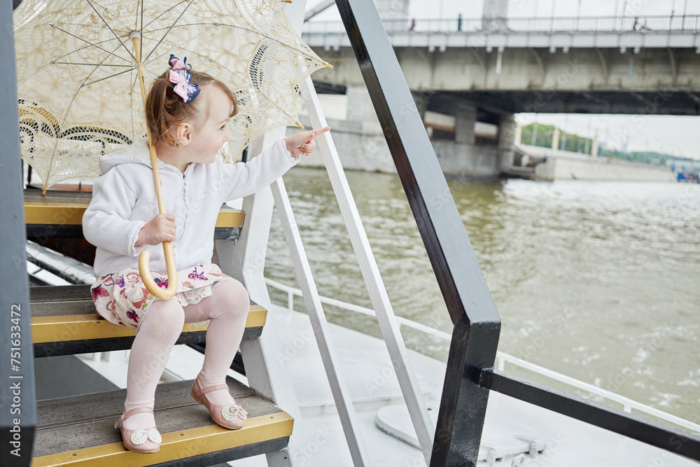 Little girl with umbrella sits on step at pleasure boat deck and points by finger - obrazy, fototapety, plakaty 