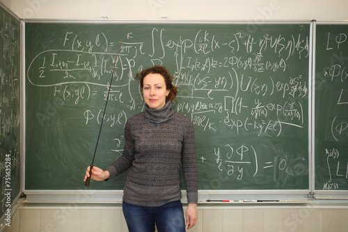 Teacher with pointer stands near blackboard with mathematics formulas in school classroom