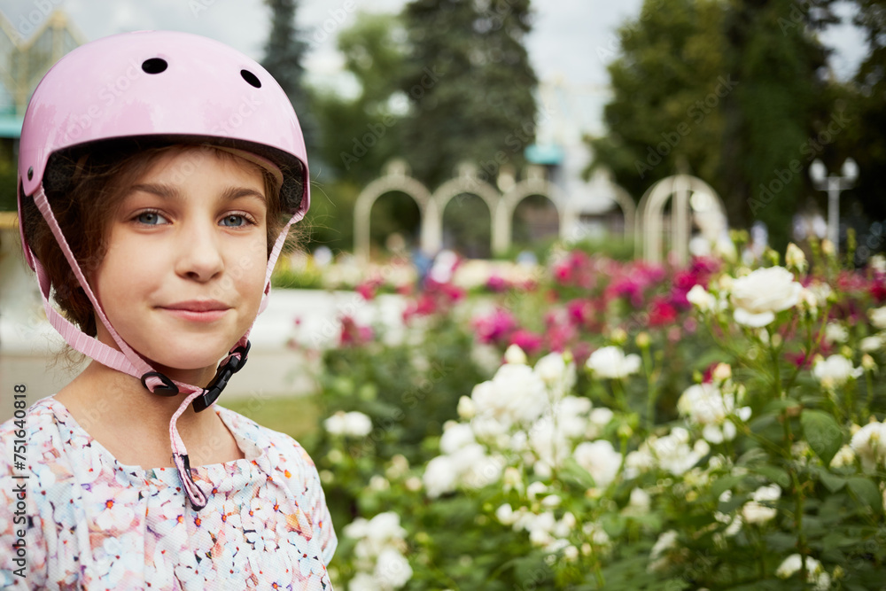 Little girl in protective pink helmet stands against flowerbed, humeral portrait Little girl in protective pink helmet stands against flowerbed, humeral portrait