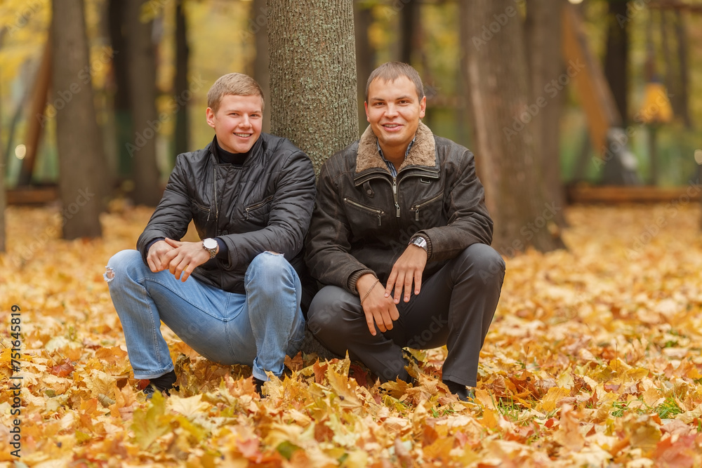 Two young man in black jacket in autumn park, crouched near tree, smiling Two young man in black jacket in autumn park, crouched near tree, smiling