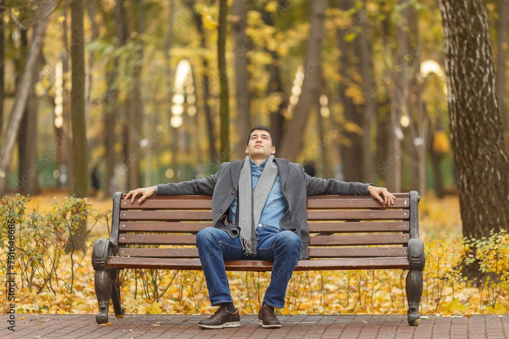 Fototapeta premium Portrait of young man in gray coat and jeans sitting on bench in alley in autumn park, hands on back, looking up
