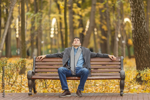 Portrait of young man in gray coat and jeans sitting on bench in alley in autumn park, hands on back, looking up