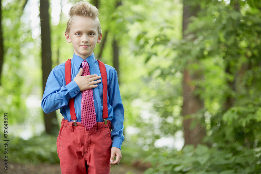 Portrait of boy in red trousers with braces and blue shirt with his hand on chest in summer park Portrait of boy in red trousers with braces and blue shirt with his hand on chest in summer park