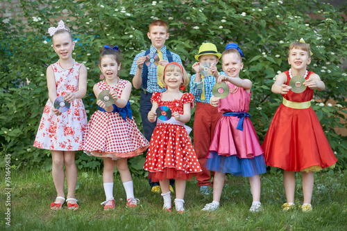 Five smiling little girls and two boys dressed in dancing suits pose at grassy lawn with cd in hands