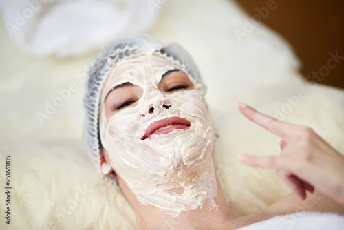 Smiling woman lying on couch in beauty salon with cosmetic mask applied on her face and nech shows goat with her left hand. photo