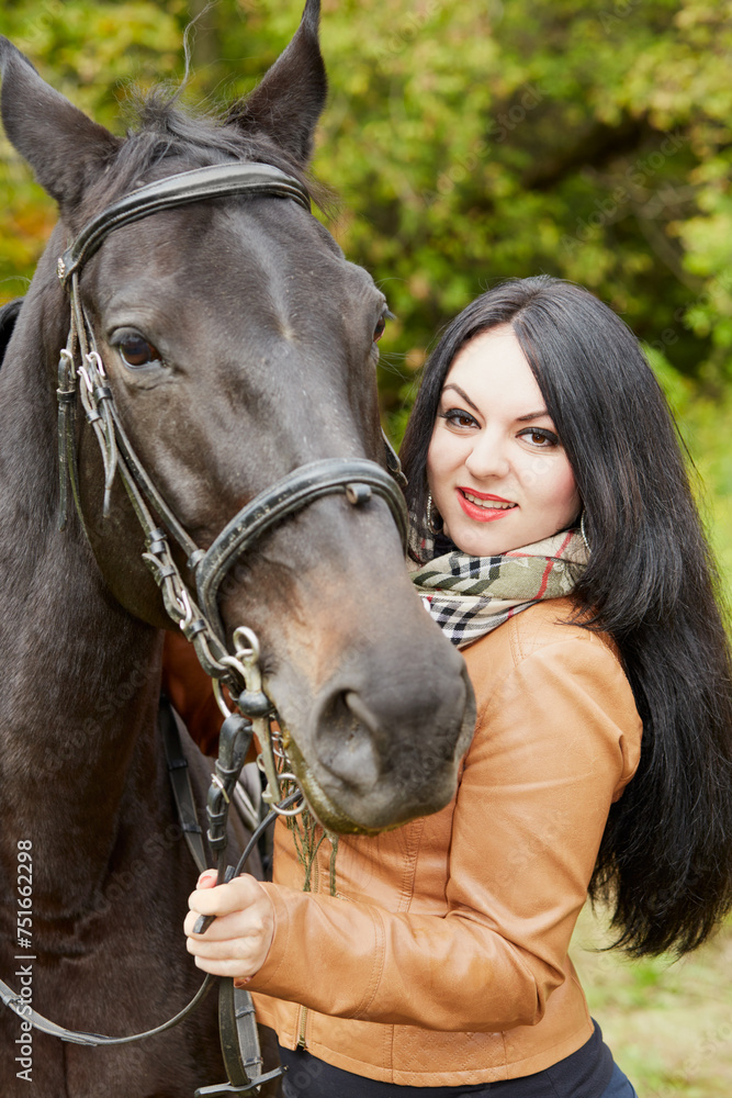 Obraz premium Smiling black-haired woman stands with horse in park.
