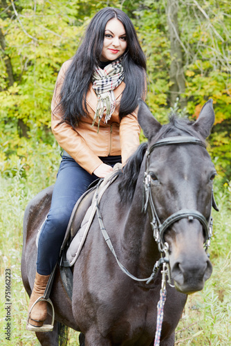 Black-haired woman in brown jacket sits on bay horse in park. © Pavel Losevsky