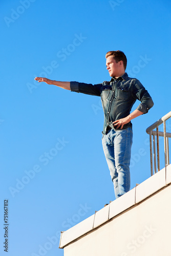 Young man in denim wear stands on the edge of the building roof outstretching one arm in front of him