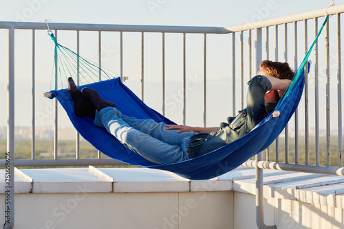 Young man in denim clothes naps in hammock attached to building roof fencing