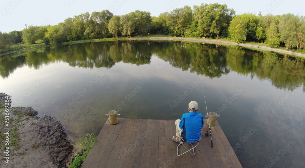 Fototapeta premium The boy with a fishing pole sitting on a wooden pier, aerial view