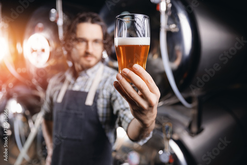 Expert brewer in apron holds glass of craft beer and checking quality and color. Worker man sommeliers taste drink on brewery factory