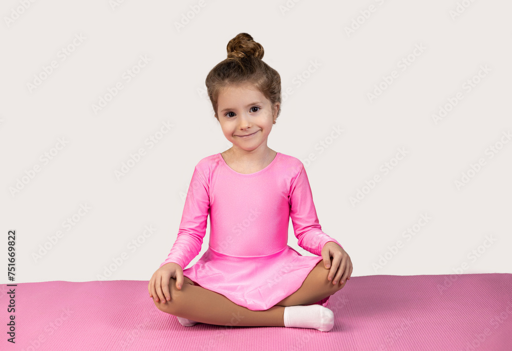 Little blonde girl in pink dress sitting on a fitness mat with her legs crossed and looking at the camera with a smile on a white background. Little blonde girl in pink dress sitting on a fitness mat with her legs crossed and looking at the camera with a smile on a white background.