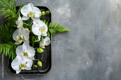 A composition of white orchids and green ferns, placed in a metal tray on a gray wall.