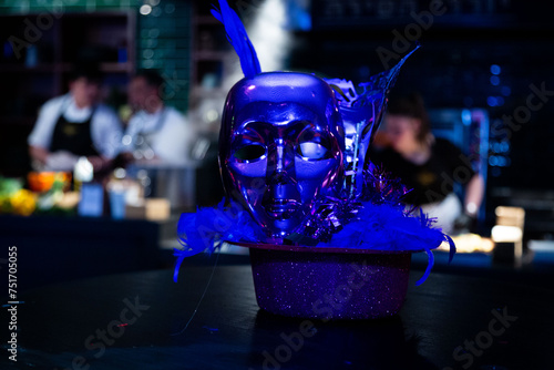 Striking blue feathered mask on glittery surface with blurred chefs in background, invoking a culinary event ambiance photo