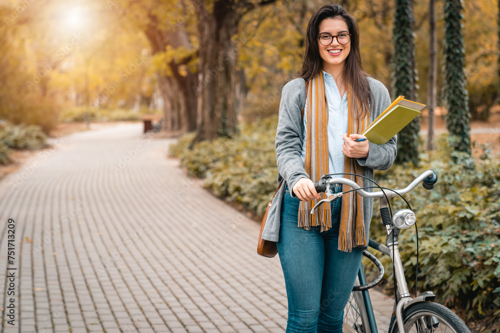 Young smiling female student with eyeglasses holding books while walking with bike in public park and smiling at camera. Young smiling female student with eyeglasses holding books while walking with bike in public park and smiling at camera.