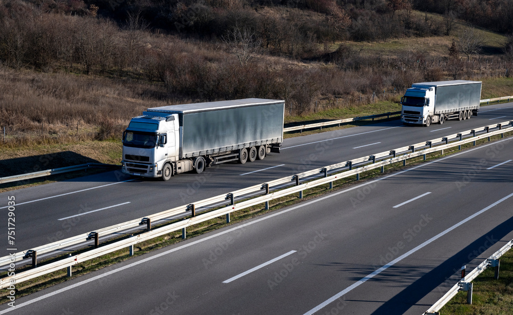 Two large trucks with gray trailers are driving on a highway through a rural area. Winter period without snow. Brown color prevails in the forest above the road.
