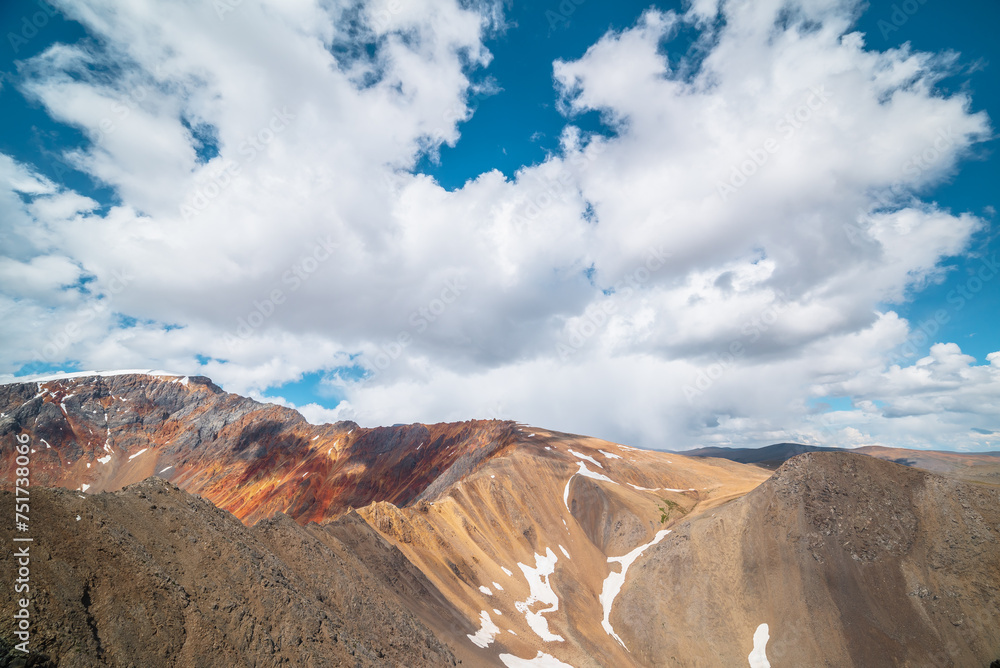 Colorful landscape with sunlit pass and sharp rocky mountain of vivid colors under cloudy blue sky. Rocks and sheer crags in sunlight. Snow and shadows of clouds on unusual multicolored mountains.