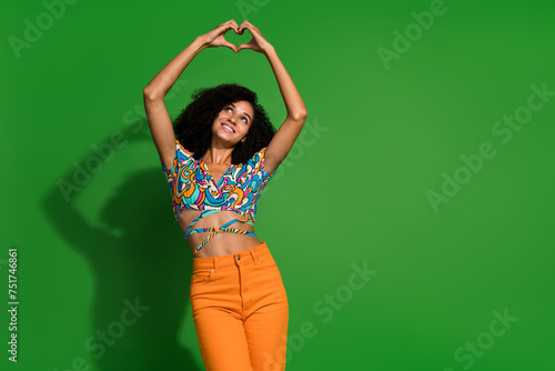 Photo of nice young woman curly hair make heart gesture looking up head cute surprise for anniversary isolated on green color background © deagreez