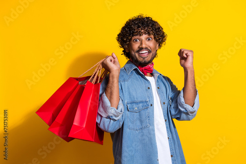 Photo portrait of attractive young man raise fist shopping bags dressed stylish denim clothes red scarf isolated on yellow color background