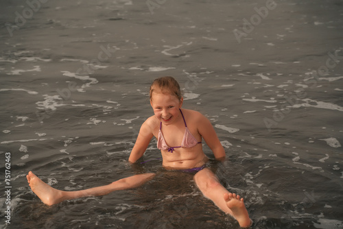 A young girl in a purple bikini laughs as she sits in the shallow seashore waters  waves lapping around. Girl Laughing in Gentle Seashore Waters