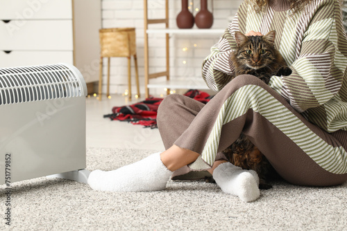 Woman with cute cat sitting on floor near electric heater at home
