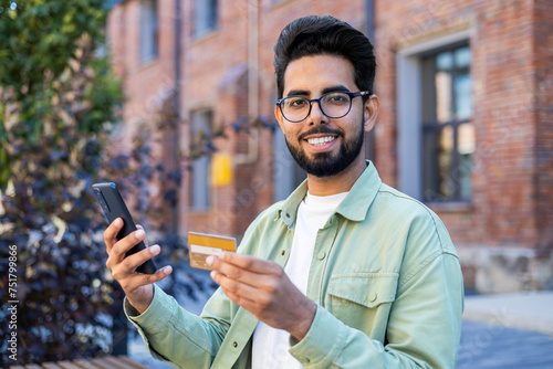 Young arab man holding mobile phone and credit card while looking at camera with wide smile. Cheery bearded guy confirming online cashless payment in banking application during walk outside.