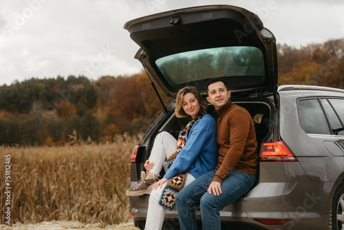 Happy young woman and man sitting in the open trunk of a car while traveling in autumn and looking away, road trip concept © Romvy