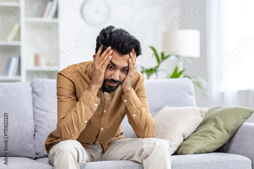 Depressed indian guy holding head with hands while staring at floor and experiencing emotional breakdown. Burnout male struggling with result of overworking while trying focusing and calming down.