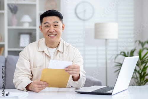 Rejoiced japanese entrepreneur receiving postal letter and reading it while working by desk with pc. Successful startup owner receiving bank notification with loan approval for business developing.