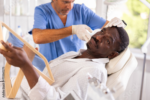 Adult dark-skinned man looking in mirror with interest while skilled male cosmetologist showing results of hardware facial procedure in office of aesthetic medicine clinic