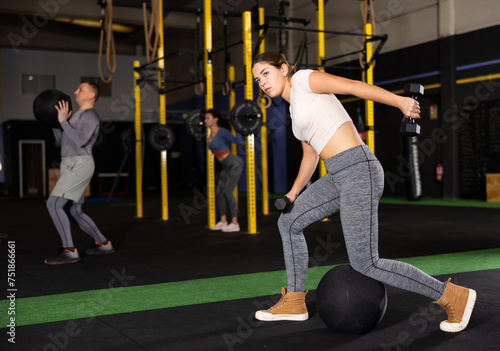 Young woman during grueling workout with dumb-bell in sports hall of fitness club. Intense exercise, increased endurance. Group and personal training, development of individual couching programs