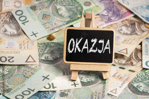 Black writing board on a wooden frame with the inscription "Okazja", Polish zloty PLN banknotes scattered in the background (selective focus) translation: chance