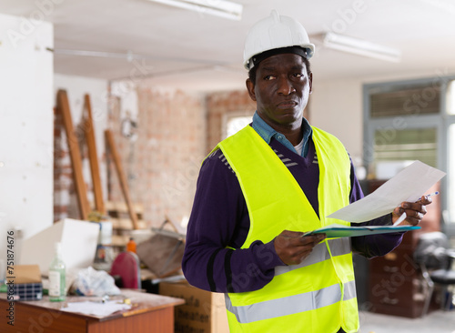 Responsible african american civil engineer conducting inspection of construction site, standing with papers indoors checking adherence to plans and blueprints