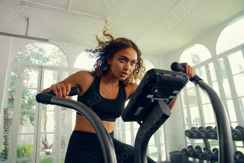 Fotografie Focused young multiracial woman in sports clothing cycling on exercise bike at t