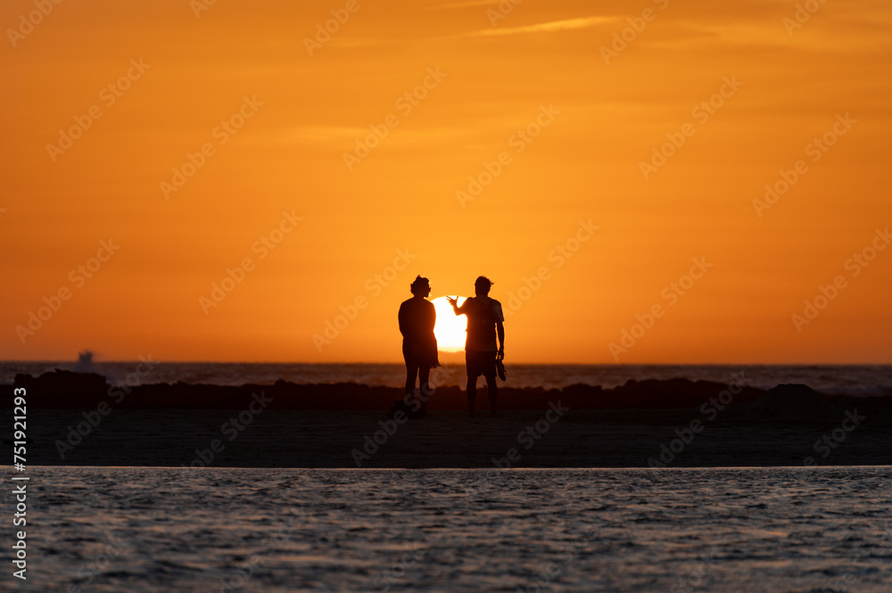 Fototapeta premium Man and woman staying and looking at sunset on ocean beach, orange sky, silhouettes of people on vacation