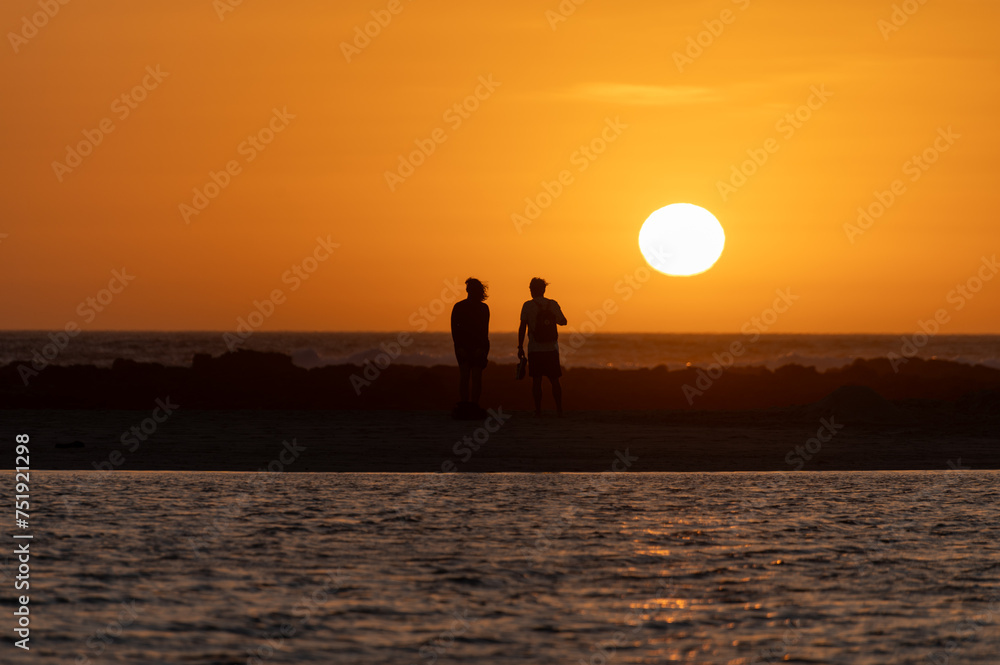 Man and woman staying and looking at sunset on ocean beach, orange sky, silhouettes of people on vacation