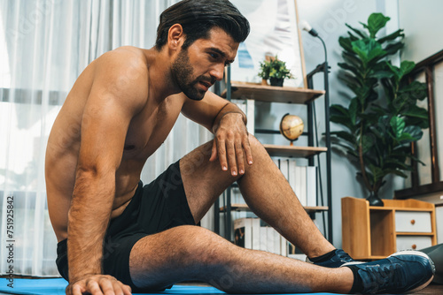 Athletic and sporty man sitting, resting on fitness mat after finishing home body workout exercise session for fit physique and healthy sport lifestyle at home. Gaiety home exercise workout training.