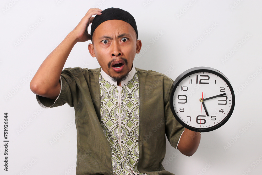 A shocked Indonesian Muslim man in koko and peci holds his head in disbelief, having realized he's late for his sahur meal during Ramadan. He holds a clock. Isolated on a white background A shocked Indonesian Muslim man in koko and peci holds his head in disbelief, having realized he's late for his sahur meal during Ramadan. He holds a clock. Isolated on a white background