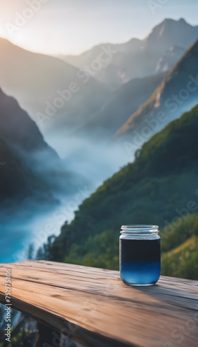 Clear jar with blue liquid on wooden surface against misty mountain landscape during sunrise.