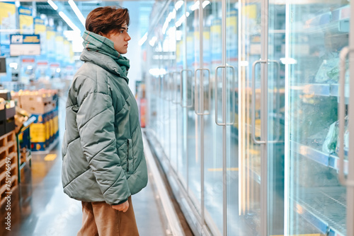 Side view of young woman shopper choosing food in refrigerator in supermarket